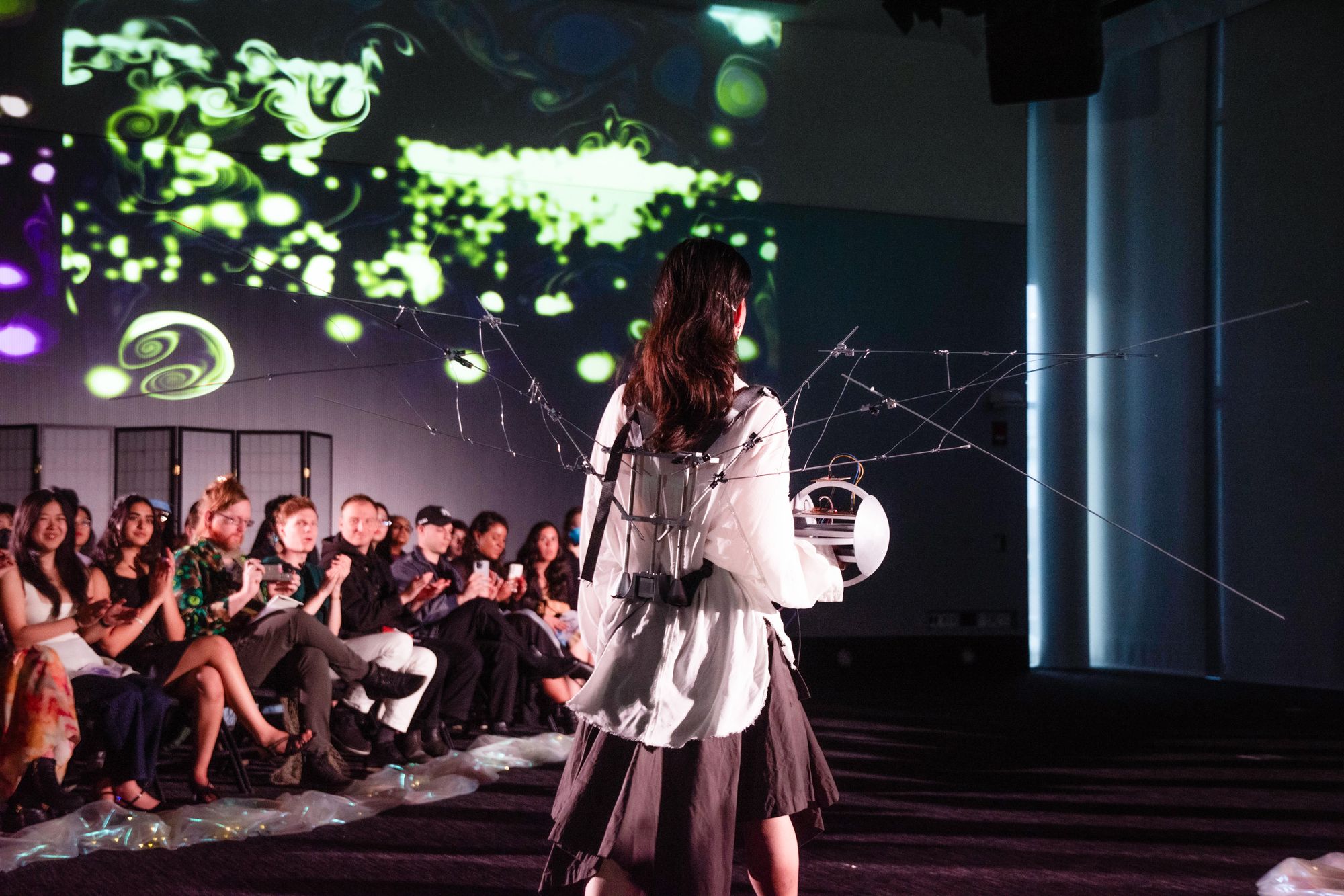A model walks the runway in front of a seated audience, wearing a wearable kinetic structure with articulated rods extending outward like wings. The apparatus is strapped to their back and lit by projections on the wall behind.
